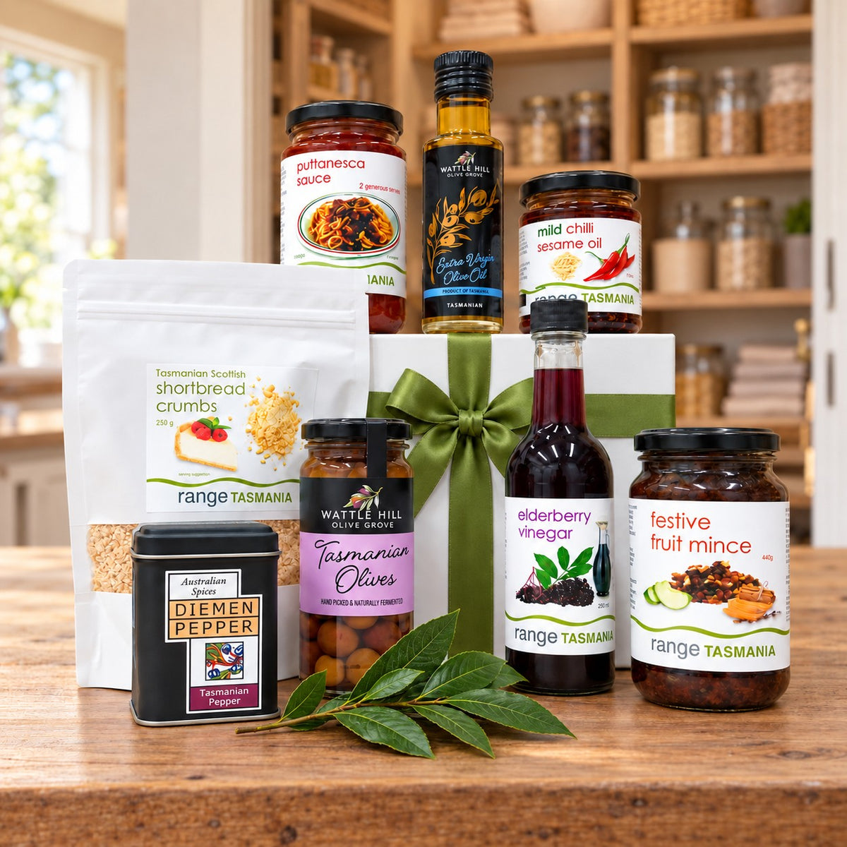 A curated gift hamper of Tasmanian food products arranged on a wooden table in front of a white gift box with a green ribbon. Visible items include jars of jam, honey, vinegar, and minces, a bag of shortbread crumbs, a tin of diemen pepper, and a bottle of elderberry vinegar, with a green leaf sprig in the foreground.