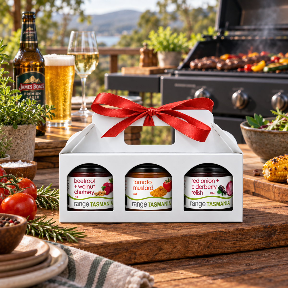 A white gift box with a red ribbon bow holds three jars from Range Tasmania, styled on a wooden table outdoors with a grill, beer, and fresh ingredients in the background.