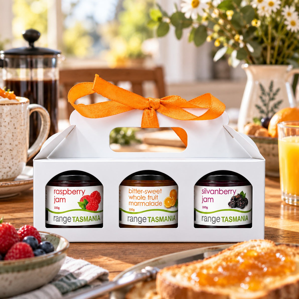 A white gift box carrying three jars of Tasmanian jam, tied with an orange ribbon, set on a breakfast table with toast, fruit, and a coffee pot in a real kitchen environment.