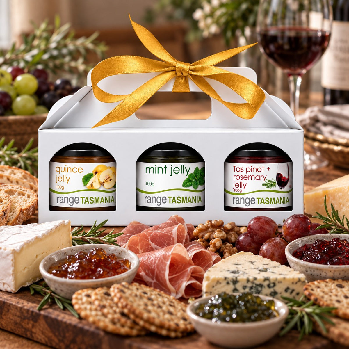 A white gift box with a gold ribbon holds three jars of Range Tasmania jelly, set on a charcuterie board with cheese, crackers, grapes, and a glass of red wine in a styled dining setting.