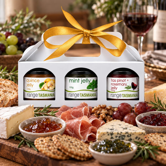 A white gift box with a gold ribbon holds three jars of Range Tasmania jelly, set on a charcuterie board with cheese, crackers, grapes, and a glass of red wine in a styled dining setting.