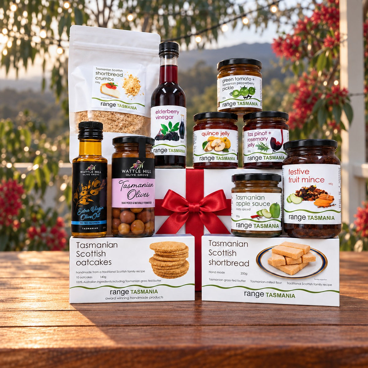 A curated gourmet gift hamper from Range Tasmania arranged on a wooden table outdoors. The gift set includes Tasmanian Scottish oatcakes in a box, shortbread cookies in a bag, strawberry vinegar, olive oil, jam, apple sauce, and nut mince, tied together with a large red ribbon.