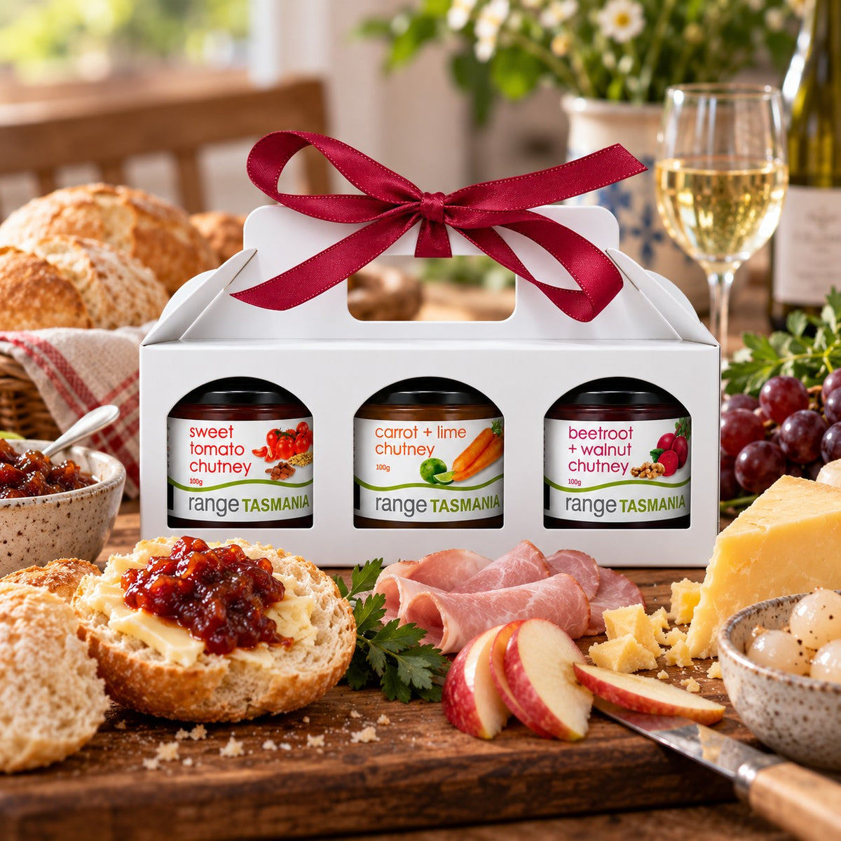 A white gift box holding three small jars of Range Tasmania spreads, tied with a dark red ribbon, displayed on a wooden table with bread, cheese, fruit, and a glass of white wine in a kitchen setting.