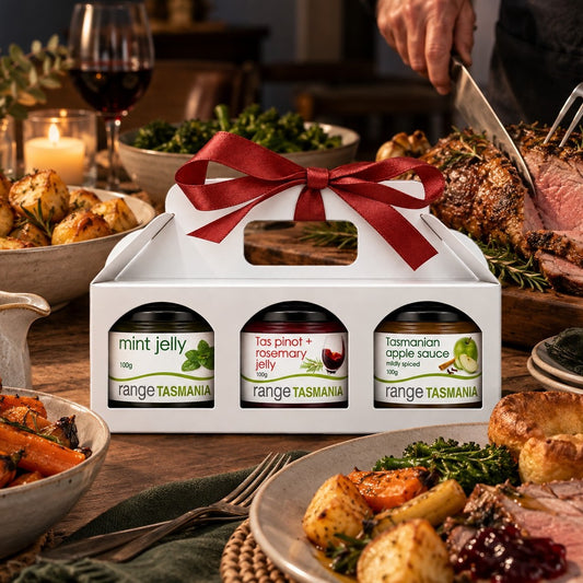 A white gift box tied with a red ribbon holds three jars of Range Tasmania spreads: mint jelly, tas pinot + rosemary jelly, and tasmanian apple sauce, set on a dining table with roasted meat, potatoes, and wine in the background.