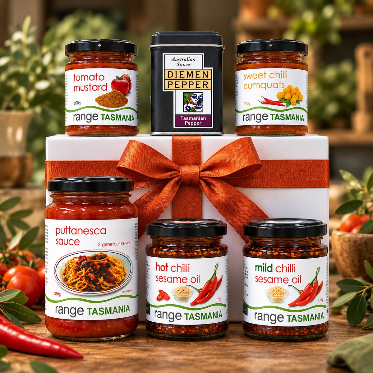 A gift set of five Range Tasmania chili and sauce products arranged around a white gift box with an orange ribbon. The set includes tomato mustard, sweet chili cumquats, puttanasca sauce, hot chili sesame oil, mild sesame oil, and a tin of Diemen Pepper.