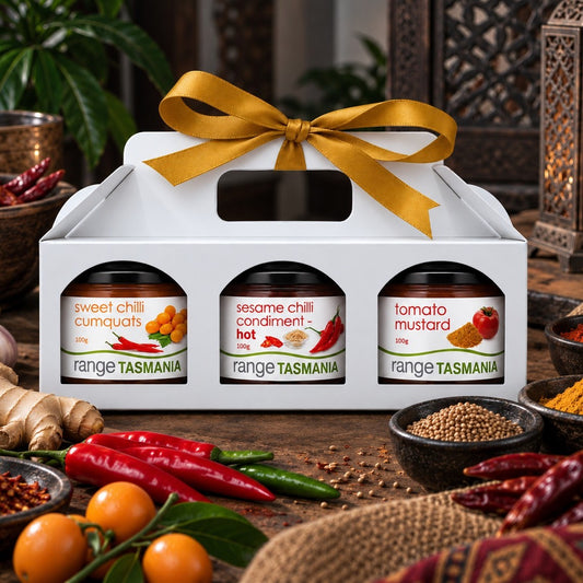 A white gift box with a gold ribbon holds three jars of Range Tasmania spices: sweet chili cumquats, sesame chili condiment hot, and tomato mustard. The box is styled on a rustic wooden table with red chilies, citrus, ginger, and bowls of spices.