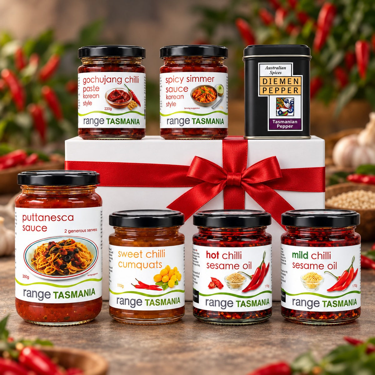 A gift set of six Range Tasmania chili sauces and spices arranged around a white gift box with a red ribbon. The set includes Puttanasca sauce, Sweet Chilli Cumquats, Hot Chilli Sesame Oil, Mild Chilli Sesame Oil, Gochujang Chili Paste, Spicy Simmer Sauce, and a tin of Diemen Pepper.