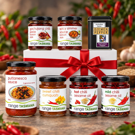 A gift set of six Range Tasmania chili sauces and spices arranged around a white gift box with a red ribbon. The set includes Puttanasca sauce, Sweet Chilli Cumquats, Hot Chilli Sesame Oil, Mild Chilli Sesame Oil, Gochujang Chili Paste, Spicy Simmer Sauce, and a tin of Diemen Pepper.